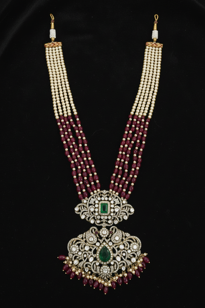 A traditional Indian Kundan necklace featuring a large central pendant with a green emerald-cut stone, surrounded by silver-toned filigree, white stones, and red beads, with three strands of cream beads transitioning to red beads, set against a black background.